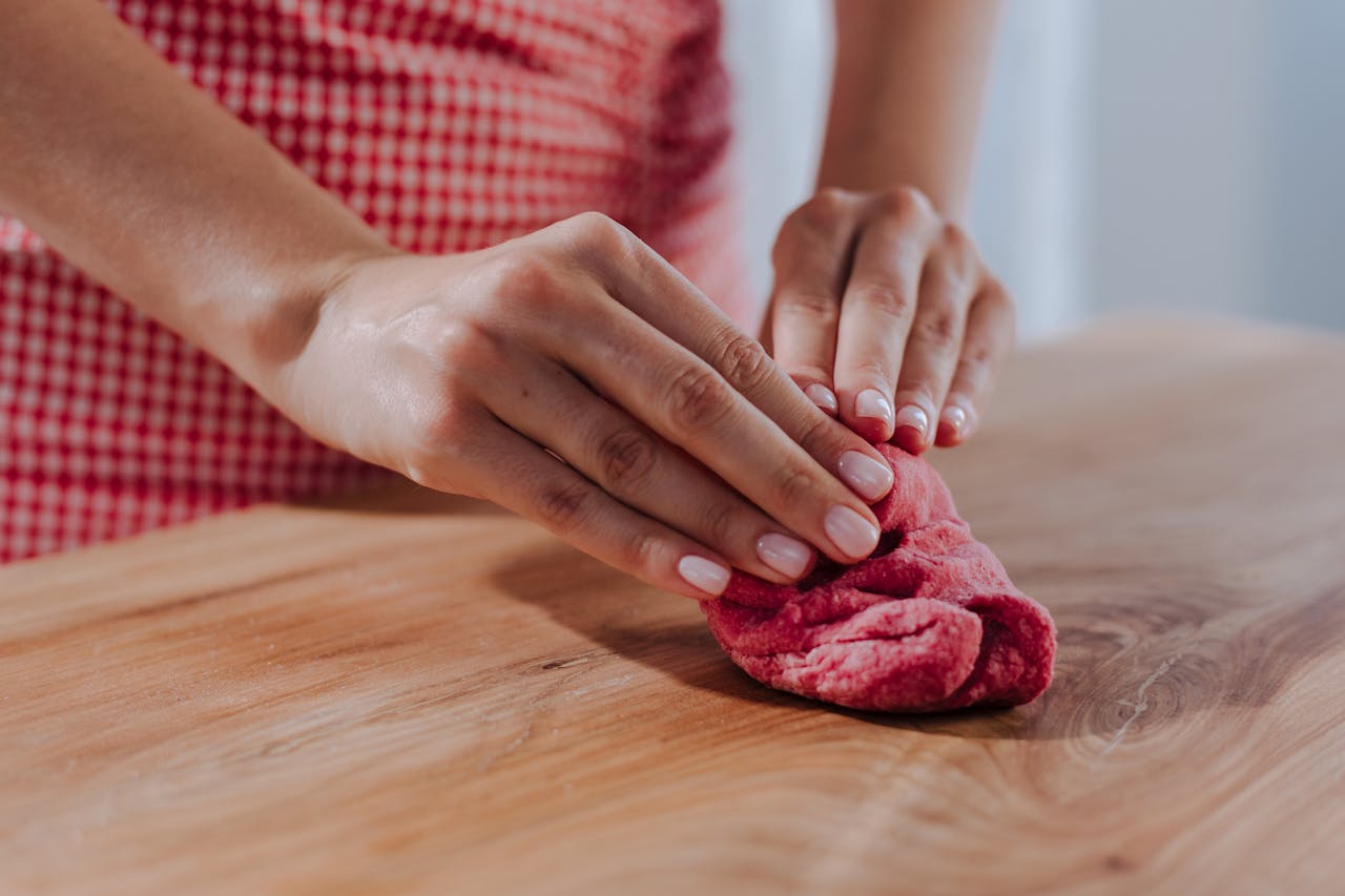 Hands kneading red dough on a wooden surface, showcasing pasta making or baking process.