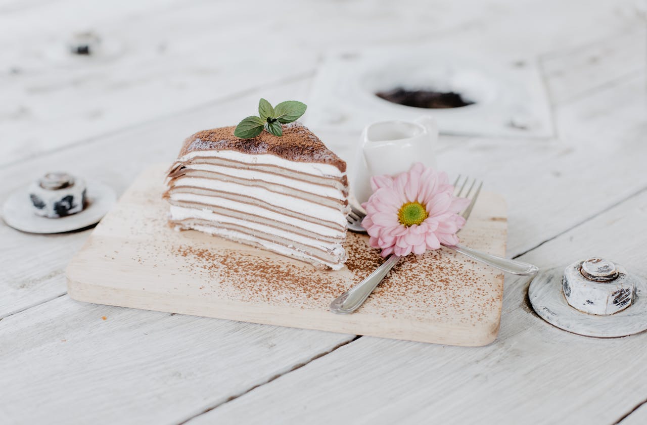 A delicate layered crepe cake garnished with mint and a pink flower on a wooden board.