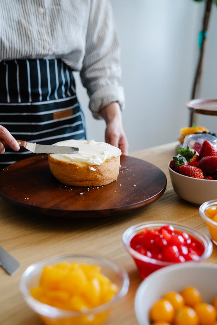 A baker decorates a vanilla cake with cream and fresh fruit in a bright kitchen setting.