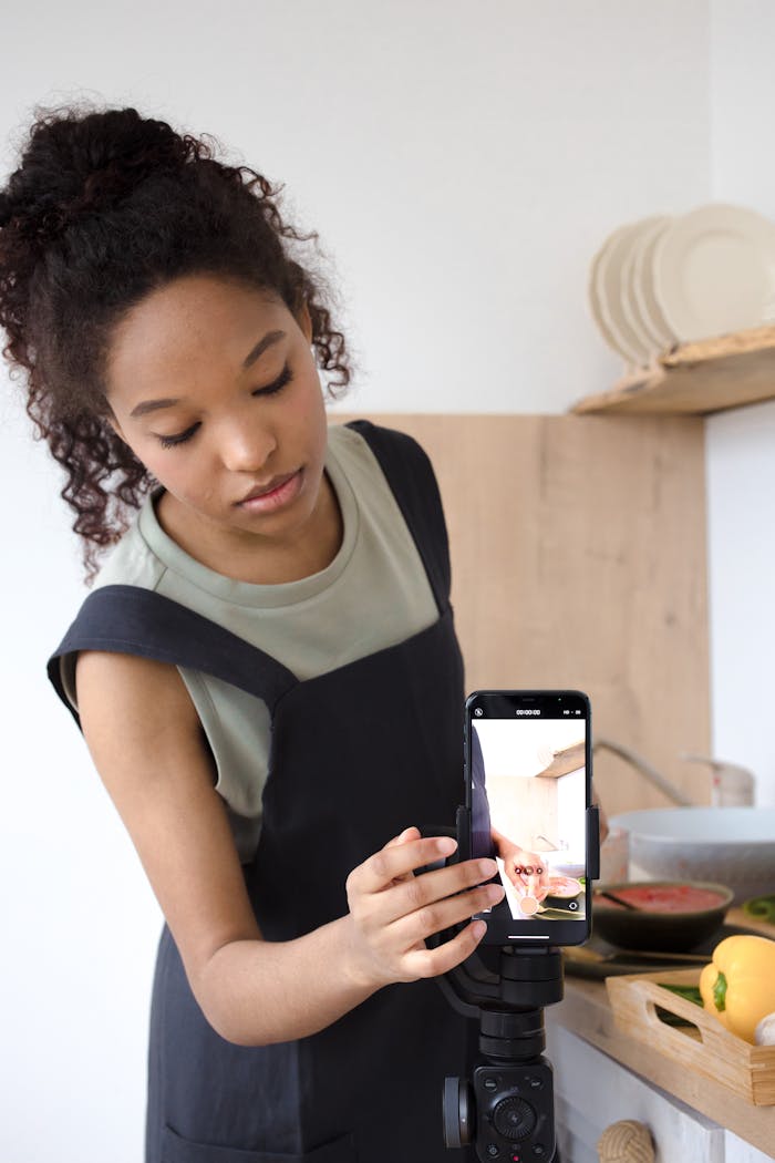 A young woman with curly hair films a cooking tutorial on a smartphone in a modern kitchen.