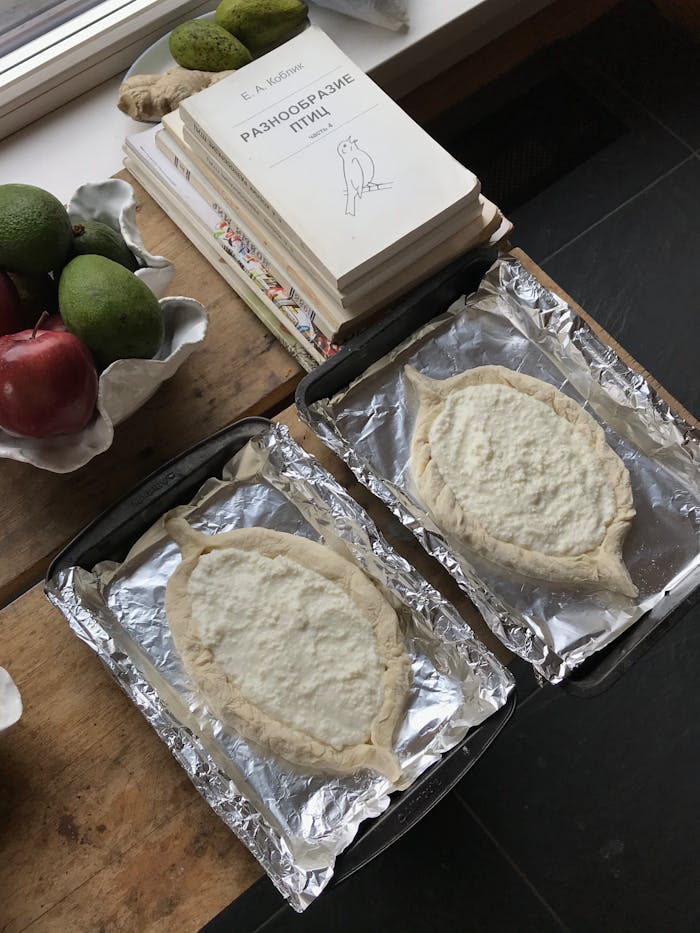 Mastering the First Impression: Your intriguing post title goes here Overhead view of raw Khachapuri on baking trays beside fresh produce and books.